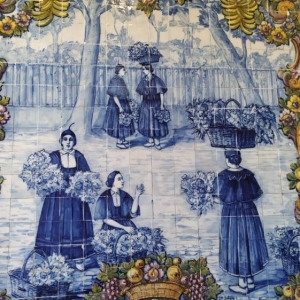 Zdjęcie przedstawia kafle powieszone na ścianie, na których jest namalowany wzór z 5 kobietami niosącymi owoce. Wokół kafli wykonany jest bardzo dekoracyjny ornament z bananami, jabłkami i gruszkami oraz herbami.
