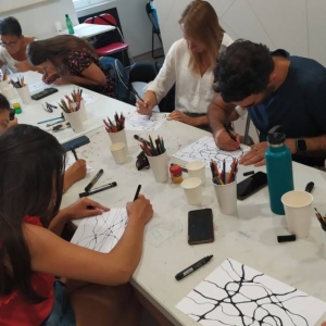 Zdjęcie przedstawia grupę osób dorosłych siedzących w pomieszczeniu przy stole. Każdy rysuje wzory czarnymi markerami na kartkach papieru.