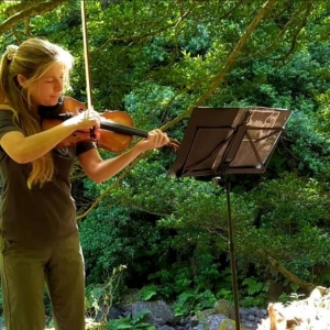 Na zdjęciu w leśnym otoczeniu stoi młoda kobieta - skrzypaczka, która właśnie gra na swoim instrumencie.