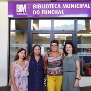 Zdjęcie przedstawia 4 kobiety stojące na zewnątrz przed budynkiem. Z tyłu widoczne są nadruki na szybach z wizerunkiem książek, a nad ich głowami zawieszony jest szyld z napisem "Biblioteca Minicipal Do Funchal" i logiem.