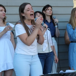 Zbliżenie na występującą na scenie młodą kobietę, która z przejęciem i zaangażowaniem śpiewa przez mikrofon. Za nią stoi czwórka kobiet, które akompaniują solistce. 
