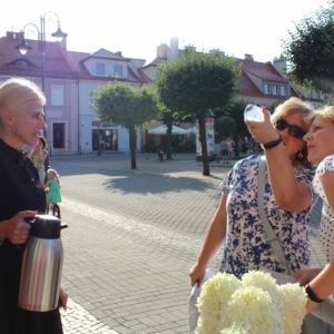 Fragment rynku w Żorach. Przy kobiecie z upiętymi włosami w kok trzymającą w rękach termos stoją dwie kobiety i spoglądają na spód szklaneczki. W tle stoją kamienice, latarnie, niewysokie drzewka.