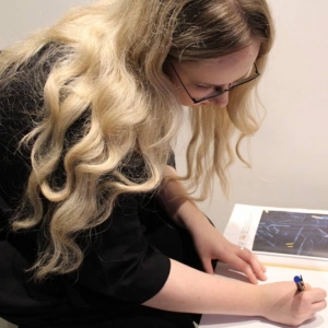 Zbliżenie na młodą kobietę, która wpisuje się do kroniki. Ma długie włosy które opadają jej na ramiona są lekko kręcone. W kronice wklejona jest kartka z grafiką i nazwą wystawy "Materia".  