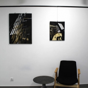 Fragment pomieszczenia. Na ścianie pod oświetleniem wystawienniczym zawieszone są dwa obrazy przedstawiające abstrakcyjne spojrzenie na architekturę. Pod ścianą stoi fotel oraz okrągły, niewysoki stolik. 