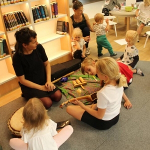 Fragment Strefy Dzieci. Na dywanie siedzą, chodzą dzieci w różnym wieku. Przy regale z książkami siedzi kobieta, obok niej leżą na dywanie instrumenty. Przed kobietą siedzi dziewczynka i gra na ukulele, a druga gra na bębnie. 