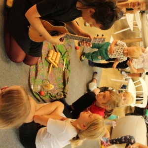 Fragment Strefy Dzieci. Na dywanie na tle półki z książkami siedzi kobieta i gra ukulele. Obok niej siedzą dzieci i patrzą na nią. Na dywanie ma ułożone inne instrumnety.