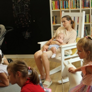 Fragment Strefy Dzieci. Na dywanie siedzą dzieci w różnym wieku. Na bujanym fotelu siedzi kobieta i trzyma na rekach dziecko. W tle na ścianie jest czarna szafla oraz regał z książkami.