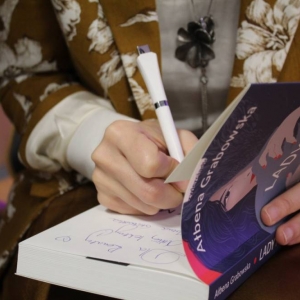 Zbliżenia na kobiece dłonie wpisujące dedykację do książki. Kobieta trzyma długopis w prawej ręce. Druga otwiera książkę. Widoczna jest okładka książki, na której jest napisane Ałbena Grabowska, tytuł Lady M. oraz grafika kobiety.