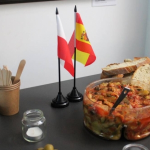 Fragment stołu, na którym wyeksponowane jest jedzenie. Chleb, warzywna sałatka, pomidory, papierowe talerzyki, serwetki, kubeczki ze sztućcami, dwie flagi Polski i Hiszpanii. 