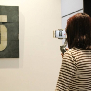 Fragment galerii. Na ścianie zawieszony jest obraz z cyfrą pięć. Przed nim stoi młoda kobieta około 20-lat i filmuje telefonem uwieszonym na gimbalu. Ma bluzkę w paski i włosy do ramion. 