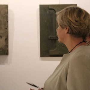 Dwie kobiety po 60-stce stoją przed obrazami zawieszonymi na ścianie.  Obrazy mają różną fakturę, na jednym jest litera I, a na drugim dziury i zniszczona celowo powierzchnia. 