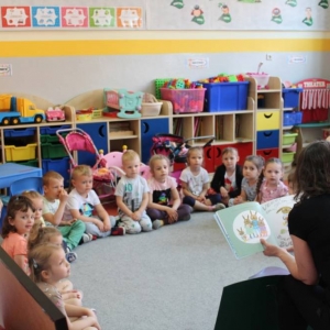 Fragment sali w przedszkolu. Pod mebelkami w przedszkolu wypełnionymi zabawkami, klockami, siedzi grupka dzieci trzyletnich i patrzy na osobę siedzącą  na krzesełku, która czyta książeczkę. Ubrana jest na czarno. 