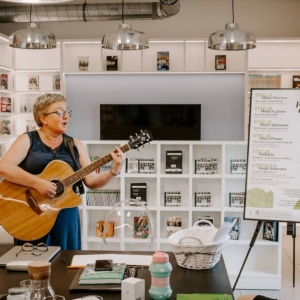W pomieszczeniu biblioteki, kobieta stoi przy stole, gra i śpiewa na gitarze. W tle widoczny jest plakat promujący projekt "Pociąg do mowy 3".