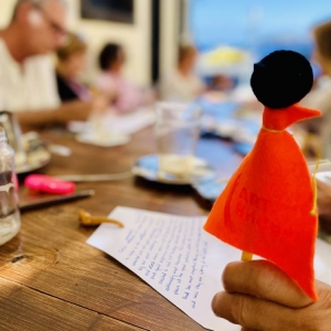 Zbliżenie na małą figurkę na ołówku. To fragment postaci w pomarańczowym płaszczu i czarnej czapce, którą trzyma kobieca dłoń. Dłoń opiera się na drewnianym stole. W tle widać zapisaną kartkę papieru oraz rozmazaną postać mężczyzny.