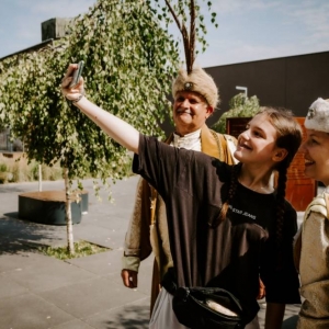 Zdjęcie przedstawia 3 dorosłe osoby. Mężczyzna i kobieta ubrani są w stroje szlacheckie. Młodsza kobieta robi sobie z nimi selfie. W tle widoczne są drzewa, budynki i kilka samochodów. 