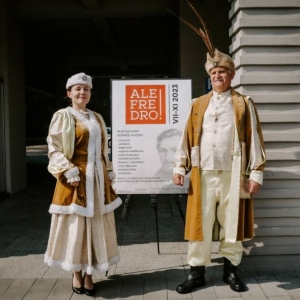 Zdjęcie kobiety i mężczyzny w strojach szlacheckich. Za nimi stoi plakat z dużym napisem "ALE Fredro VII-XI".