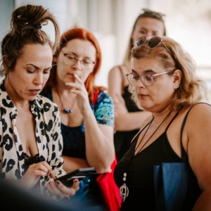 Na zdjęciu widoczne są 4 kobiety w średnim wieku. Jedna z nich, będąca najbliżej fotografa ma upiętego wysoko koka, bluzkę z dość dużym dekoldem i trzyma w dłoniach telefon, na którym coś pokazuje. Pozostałe kobiety patrzą na jej telefon.