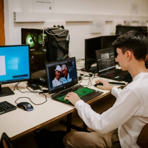 Wnętrze pracowni komputerowej. Zdjęcie przedstawia zbliżenie na jednego z uczniów. To około 20 letni chłopak, który wpatruje się w monitor swojego laptopa. Widać, że jest skupiony na wykonywanej pracy. W tle widoczni są pozostali uczniowie i komputery.