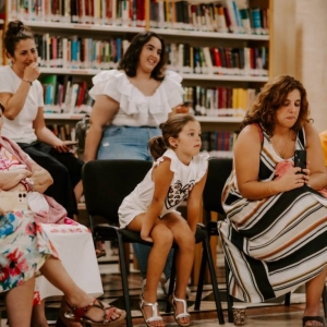 Zbliżenie na grupę ośmiu osób siedzących na krzesłach w bibliotece. W grupie tej dominują kobiety w różnym wieku, jedyny mężczyzna siedzi po prawej stronie i dość mocno wyróżnia się jaskrawym kolorem swojej koszulki. Kobieta siedząca obok niego trzyma przed sobą telefon i wykonuje fotografię. Wszyscy wpatrują się przed siebie. Za nimi widoczne są regały z książkami.