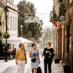 Ujęcie grupy trzech rozmawiających ze sobą osób, które przemierzają uliczkę starego miasta. Wszyscy są w wieku około 65 lat. Po lewej idzie mężczyzna w jasnych spodniach i żółtej koszuli. Ma półdługie, siwe i kręcone włosy. Spogląda na kobietę po jego lewej. Kobieta ma jasnoniebieską, dopasowaną sukienkę i czarną, małą torebkę w jednej dłoni trzyma małe pudełko, a w drugiej czarny plecak. Spogląda na kobietę po prawej stronie. Ma ona ciemne spodnie i bluzkę oraz okulary przeciwsłoneczne.