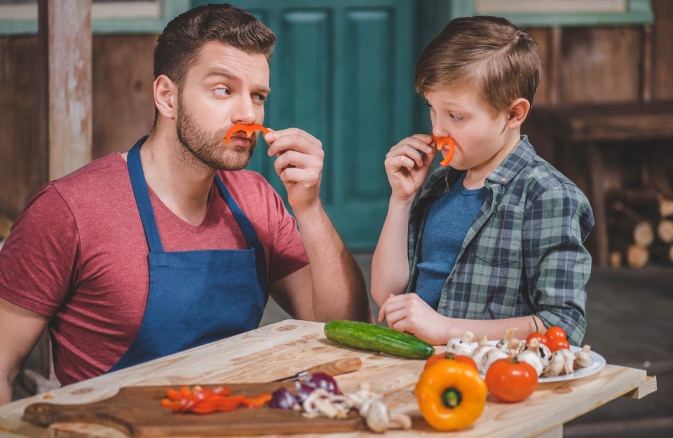Zdjęcie ojca z synem. Kroją różne warzywa,a  przy ustach trzymają kawałki papryki wyglądające jak wąsy.